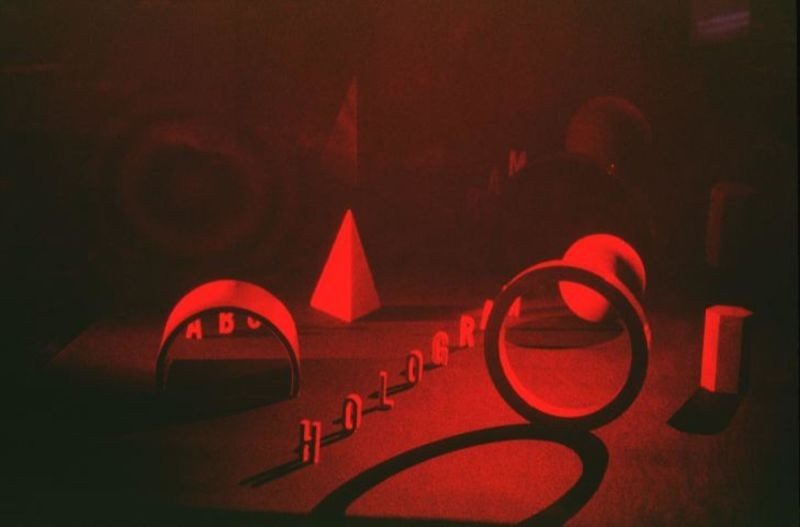 Series of geometric objects (cones, cubes, hoops, circles, etc.) set about a board; the word `HOLOGRAM,' made out of wooden stand-up letters, stretches diagonally into background; good, early display of 3-D effect of holography; red image. Photo provided by the MIT Museum.