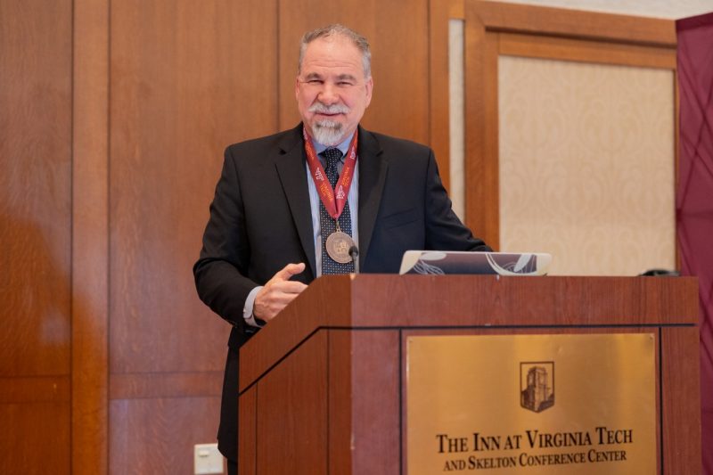 Tom Drayer, wearing a medal around his neck, stands behind a podium at the Inn at Virginia Tech.